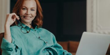 Indoor shot of pleasant looking redhead woman has good time at home, works on computer