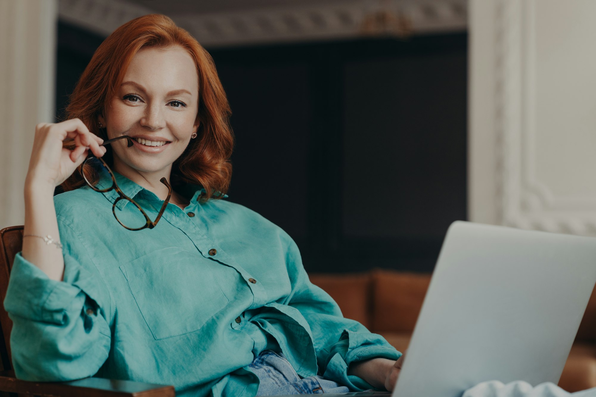 Indoor shot of pleasant looking redhead woman has good time at home, works on computer