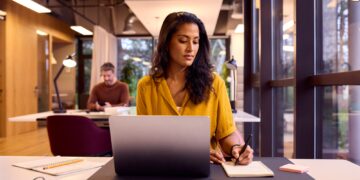 Mature Businesswoman Working On Laptop At Desk In Office Writing In Notebook